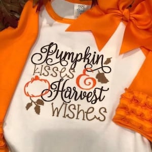 May include: White and orange long-sleeved shirt with the text "Pumpkin kisses Harvest wishes" in brown script. The shirt has an orange bow and ruffled cuffs. The shirt is decorated with pumpkin and leaf designs.