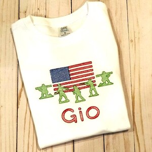 May include: White t-shirt with an embroidered American flag and a line of green toy soldiers. The name "Gio" is embroidered below the soldiers.