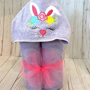 May include: A lavender hooded towel with a bunny face embroidered on the hood. The bunny has white ears, pink nose, and a pink heart. The towel is tied with a pink tulle bow.