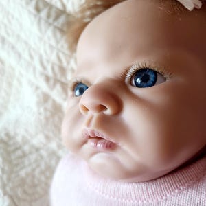 Puede incluir: Primer plano de una muñeca bebé con ojos azules y un atuendo rosa. La muñeca tiene una apariencia realista y está acostada sobre una manta blanca.