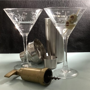 Peut inclure: Deux verres à martini en verre transparent avec un motif en étoile gravé dans le verre. Les verres sont remplis d'un liquide clair et garnis d'olives vertes. Un shaker à cocktail argenté et un jigger argenté sont également présents sur l'image.