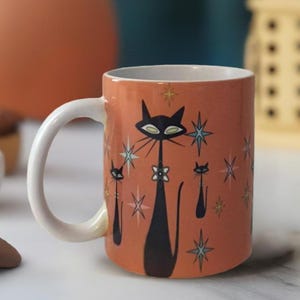 May include: A ceramic coffee mug with a retro design featuring three black cats with white eyes and a starburst pattern on an orange background.