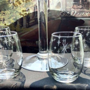 May include: A set of clear stemless wine glasses with etched star designs. The glasses are arranged on a dark surface, with a tall, clear stem in the background. The background features a blurred image of a street scene.