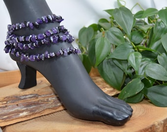 May include: Ankle bracelet featuring multiple strands of purple and silver beads. The jewellery is displayed on a black mannequin foot. The beads are small, irregular stones. The background includes wood and greenery.
