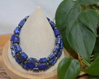 Pode incluir: Uma pulseira de duas voltas com contas retangulares de lápis-lazúli azul escuro e pequenas contas metálicas. A pulseira é exibida em um suporte bege em forma de cone, com folhagem verde ao fundo.