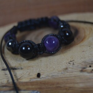 Puede incluir: Un brazalete de cordón negro con cuentas moradas y negras. El brazalete está anudado y tiene un borla.