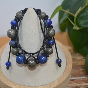 Op de afbeelding: Een armband van zwart koord met blauwe en gouden kralen. De armband is met een knoop vastgemaakt en heeft aan elk uiteinde een kwast.