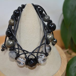 Op de afbeelding: Een zwart kralenarmband met een geweven ontwerp. De armband heeft zwarte onyx, pyriet en heldere kwartskralen. De armband wordt gepresenteerd op een beige standaard.