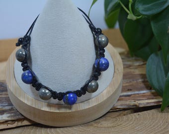 Pode incluir: Um bracelete de cordão preto com contas azuis e douradas. O bracelete é exibido em um suporte de madeira.