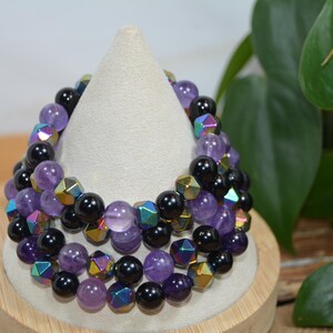 Può includere: Una pila di tre braccialetti di perline. I braccialetti presentano una combinazione di perline nere, viola e geometriche iridescenti. Sono esposti su un supporto bianco a forma di cono. Sullo sfondo si vede del fogliame verde.