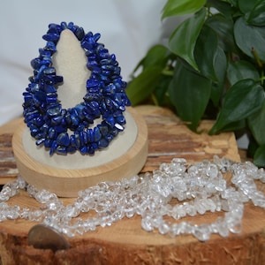 May include: A multi-strand bracelet made of deep blue lapis lazuli chip beads, displayed on a beige cone stand. Beside it lies a long, loose strand of clear, irregular quartz crystal chip beads, showcasing a natural, polished, and raw aesthetic.