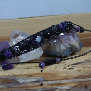 Op de afbeelding: Een handgemaakte macramé armband met amethist en zwarte kralen. De armband is geregen op een zwart koord en bevat een gefacetteerde amethiststeen. De verstelbare armband wordt gepresenteerd op een houten oppervlak met amethistkristallen.