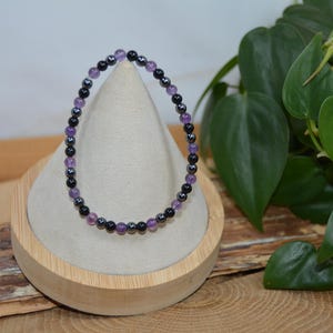 May include: A beaded bracelet featuring alternating purple and black beads. The bracelet is displayed on a wooden stand with a neutral fabric. Green leafy plants are in the background.