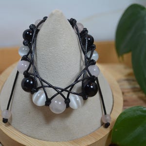 Op de afbeelding: Een handgemaakte armband met zwarte, witte en roze kralen geregen op zwart koord. De armband wordt gepresenteerd op een beige standaard, die het ingewikkelde ontwerp en de kleurencombinatie benadrukt. De kralen zijn rond en glad.