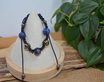 Pode incluir: Um bracelete de cordão preto com contas azuis e douradas. O bracelete é exibido em um suporte de madeira.