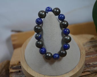 Pode incluir: Uma pulseira de contas com contas esféricas cinza escuro e azul profundo alternadas. A pulseira é exibida em um suporte em forma de cone cinza claro com uma base de madeira. O fundo inclui madeira e vegetação.