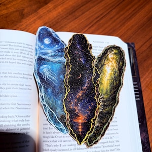 May include: A set of three irregular, leaf-shaped bookmarks with gold-painted edges. The left bookmark features a blue ocean scene with a jellyfish and moon. The center shows a starry night sky over water. The right depicts a sunlit forest path.