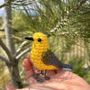 May include: A handmade crochet bird with a bright yellow body, gray wings, and a black beak. The bird has small, detailed wire legs and is set against a backdrop of green pine needles and a blue sky. The bird is approximately 3 inches tall.