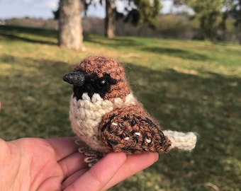 May include: A small crocheted brown and white bird with black eyes. The bird is sitting in a person's hand.