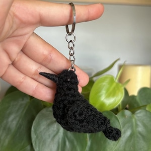 May include: Black crocheted bird keyring with a silver key ring. The bird has black bead eyes and is perched on a green plant.