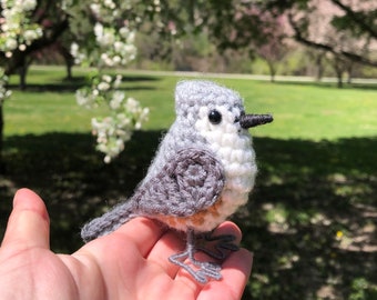 May include: A crocheted gray and white bird with black eyes and a small orange beak. The bird is sitting on a person's hand.