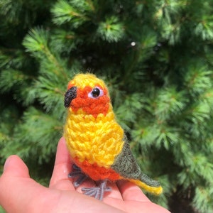 May include: A crocheted sun conure bird toy, yellow with orange and green accents. The bird has black eyes and a small black beak.