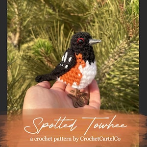 May include: A handmade crochet Spotted Towhee bird with black, white, and orange plumage. The bird has a detailed design, including a small beak and red eyes. The text "Spotted Towhee" and "a crochet pattern by CrochetCartelCo" are visible.