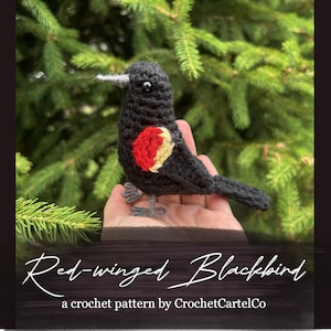 May include: A handmade crochet blackbird with a red and yellow wing patch, set against a backdrop of green leaves. The bird features a silver beak and wire legs. The text reads "Red-winged Blackbird" and "a crochet pattern by CrochetCartelCo".