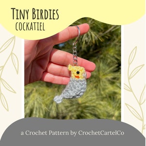 May include: A crocheted cockatiel keychain with yellow and gray yarn. The keychain is attached to a silver chain and is being held by a hand. The image has the text "Tiny Birdies Cockatiel" and "a Crochet Pattern by CrochetCartelCo".