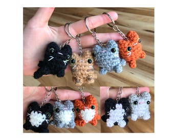 May include: A set of nine crocheted cat keychains in various colors, including black, gray, orange, and brown. Each keychain features a small crocheted cat with white facial details.