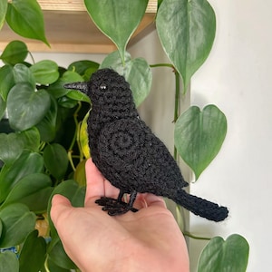 May include: A black crocheted bird with a small, round body and a long, thin tail. The bird is sitting on a person's hand.