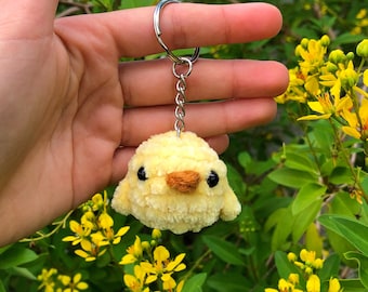 May include: A yellow fluffy chick keychain with black eyes and an orange beak. The keychain is hanging from a silver chain and ring.