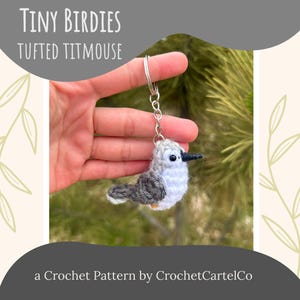May include: A crocheted Tufted Titmouse bird keychain with a silver keyring. The bird is gray, white, and black, with a small black beak. The text "Tiny Birdies Tufted Titmouse" is at the top of the image, and "a Crochet Pattern by CrochetCartelCo" is at the bottom.
