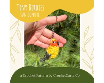 May include: A crocheted sun conure bird keychain. The bird is yellow, orange, and gray, with a silver keychain ring. The text "Tiny Birdies Sun Conure" is at the top, and "a Crochet Pattern by CrochetCartelCo" is at the bottom.