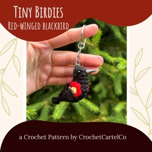 May include: A crochet keychain depicting a red-winged blackbird, with black, red, and yellow detailing. The bird has a silver beak and is attached to a silver keyring. The background is green foliage. Text reads "TINY BIRDIES Red-winged Blackbird".