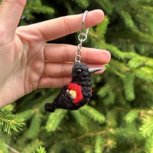 Puede incluir: Un llavero de pájaro negro hecho a mano con un acento rojo y amarillo en su costado. El pájaro tiene un pico plateado y está unido a un llavero plateado. El llavero está hecho de hilo y se muestra sobre un fondo de follaje verde.