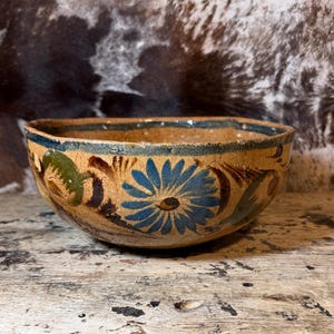 May include: A rustic, hand-painted ceramic bowl with a floral design. The bowl features a tan base with blue, green, and brown floral patterns around the exterior. The rim is painted with a dark blue band. The bowl is likely used for serving.