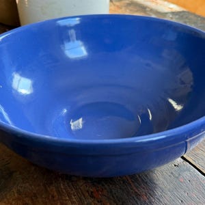 May include: A large, round, cobalt blue ceramic bowl. The bowl has a wide rim and a smooth, glossy finish. It sits on a weathered wooden surface, with a glimpse of other ceramic items in the background. The bowl is empty.