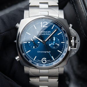 May include: A silver-toned stainless steel Luminor Panerai chronograph watch with a deep blue dial. The watch features a prominent crown guard, a tachymeter scale on the outer rim, two sub-dials, and the text Luminor Panerai Chronograph.