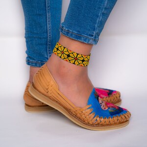 May include: A pair of tan leather sandals with a blue and pink Frida Kahlo design. The sandals have a woven strap and a yellow and black beaded ankle bracelet.