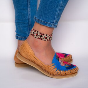May include: A pair of tan leather sandals with a woven design. The sandals feature a blue and pink print of a woman's face. The woman is wearing a flower crown. The sandals are being worn by a person with a beaded ankle bracelet.