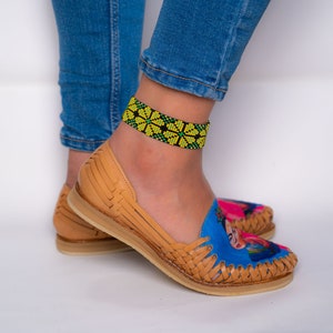 May include: A pair of tan leather sandals with a blue and pink Frida Kahlo design. The sandals have a woven strap and a yellow and green beaded ankle bracelet.