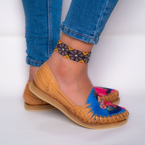 May include: A pair of tan leather sandals with a woven design. The sandals have a blue and pink Frida Kahlo print on the toe. The person wearing the sandals is also wearing a beaded ankle bracelet with black, yellow, and orange beads.