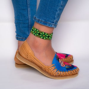 May include: A pair of tan leather sandals with a blue and pink Frida Kahlo design. The sandals have a woven strap and a green and black beaded ankle bracelet.