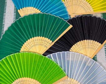 Può includere: Una collezione di ventagli aperti in vari colori, tra cui turchese, giallo, verde, nero, verde chiaro e azzurro. Ogni ventaglio presenta un manico in legno e un design a pieghe. I ventagli sono disposti su uno sfondo a righe verdi e bianche.