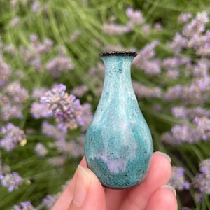 May include: A miniature ceramic vase with a bulbous body and a narrow neck, featuring a glossy glaze in shades of turquoise, teal, and hints of soft purple, with a dark brown rim and a speckled, textured finish throughout the surface.