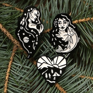 May include: Three black and white enamel pins featuring a woman with stars in her hair, a woman holding a fish, and a heart-shaped design with a mountain and sun.