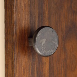 Peut inclure: Un bouton de placard circulaire en métal gris foncé avec une finition texturée et patinée, fixé sur un panneau vertical en bois brun foncé présentant un grain linéaire marqué courant du haut vers le bas de la surface.