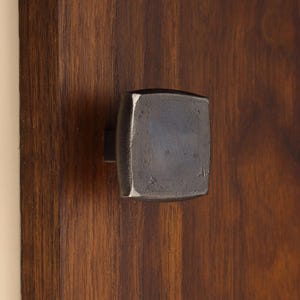May include: A square, dark grey metal cabinet knob with a slightly hammered, textured surface. The knob is mounted onto a vertical, dark brown wooden panel with a visible grain pattern, showcasing a contrast between the metal and wood.