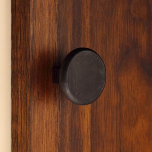 May include: A matte black circular cabinet knob attached to a dark brown wooden surface with a visible vertical grain pattern. The knob features a simple, flat-faced design with a small square base connecting it to the wood panelling.